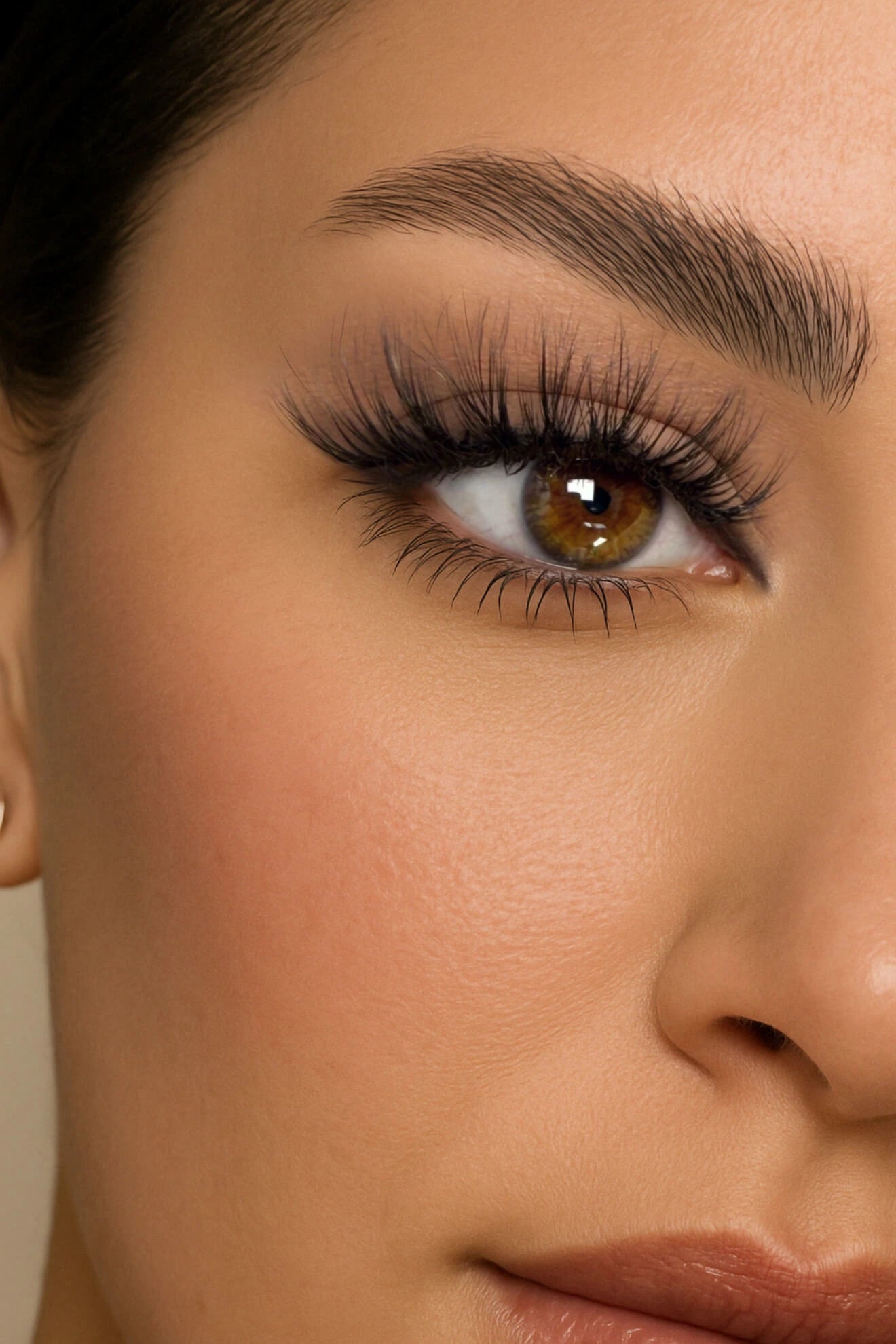 Close-up of a woman's face showcasing voluminous false eyelashes, warm-toned makeup, and a natural, glowing complexion with brown eyes.